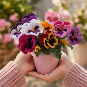 May include: A potted arrangement of crocheted pansy flowers in shades of purple, white, yellow, pink, and magenta. The flowers feature dark centers and are attached to green crocheted leaves, all contained within a solid light pink ceramic flower pot.