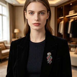 Può includere: Una spilla a forma di medusa color argento con una cupola testurizzata e tentacoli pendenti decorati con piccole pietre preziose rosse, verdi e blu. La spilla è appuntata sul bavero di un blazer in velluto nero indossato sopra una maglia nera.