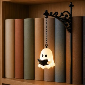Op de afbeelding: Een wit, oplichtend spookvormig lampje dat aan een dunne ketting hangt aan een zwarte, sierlijke metalen wandbeugel. Het spookfiguurtje houdt een klein, zwart open boek in zijn handen en verspreidt een zacht, warm schijnsel in de ruimte.