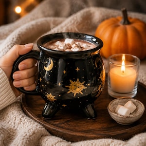 Op de afbeelding: Een zwarte keramische mok in de vorm van een ketel met gouden hemelse ontwerpen van zonnen, manen en sterren. De mok heeft drie kleine pootjes en is gevuld met een warme drank met witte marshmallows, waar zichtbare stoom vanaf komt.