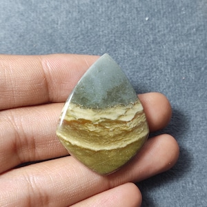 May include: A teardrop-shaped polished gemstone cabochon featuring horizontal bands of color. The top section is a muted sage green, transitioning into layers of cream, tan, and a deeper olive green at the base with a smooth, glossy finish.