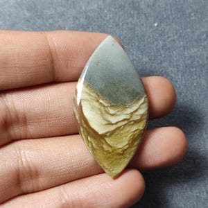 May include: A marquise-shaped polished gemstone cabochon featuring a smooth surface with a distinct color gradient. The top section is a solid muted gray, while the bottom portion displays layered, wavy bands of cream, tan, and golden-brown tones.