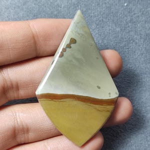 May include: A polished, kite-shaped cabochon gemstone featuring a smooth surface with distinct color bands. The top section is a muted sage green with small brown spots, while the bottom section is a solid mustard yellow, separated by a thin rust-colored line.