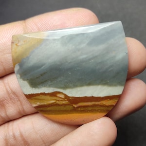May include: A polished, semi-circular cabochon gemstone featuring horizontal bands of color. The stone displays a large section of muted blue-gray at the top, followed by a thin white stripe, and layers of earthy brown, tan, and golden yellow at the base.