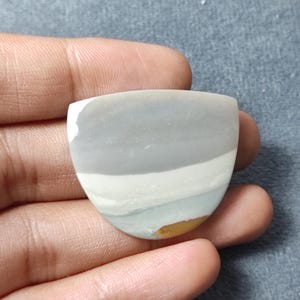 May include: A polished, shield-shaped cabochon stone featuring horizontal bands of color. The surface displays a smooth transition from a solid light gray top section to a white middle band, followed by a pale blue-green base with a small yellow spot.