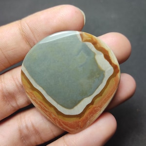 May include: A polished, teardrop-shaped cabochon stone featuring concentric bands of color. The center is a muted sage green, surrounded by distinct layers of cream, golden yellow, and a vibrant, earthy terracotta orange at the outer edge.