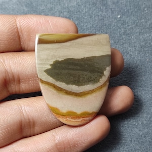 May include: A polished shield-shaped cabochon gemstone featuring horizontal bands of color including tan, olive green, mustard yellow, and cream. The stone has a smooth, flat surface with a rounded bottom edge and a straight top edge.
