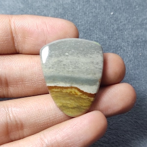 May include: A polished, shield-shaped cabochon gemstone featuring horizontal bands of color. The top section is a smooth, light gray, transitioning into a thin dark gray line, followed by a band of white, and a textured, earthy mustard yellow base.