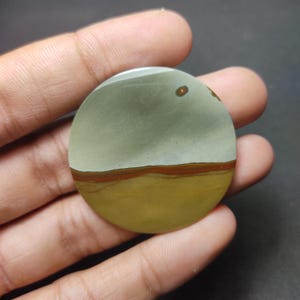 May include: A circular polished gemstone cabochon featuring a smooth surface with distinct horizontal color bands. The top section is a muted sage green, separated by a thin dark brown line from the bottom section, which is a solid mustard yellow.