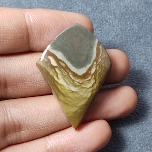 May include: A kite-shaped polished gemstone cabochon featuring a smooth surface with distinct color bands. The stone displays a top section of muted grey, transitioning into a white line, followed by layered patterns of tan, ochre, and olive green.