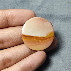 May include: A circular, polished gemstone cabochon featuring a smooth, flat surface with horizontal bands of color. The stone displays a gradient of cream, soft peach, and warm orange tones, with distinct, wavy, golden-yellow and brown layers.