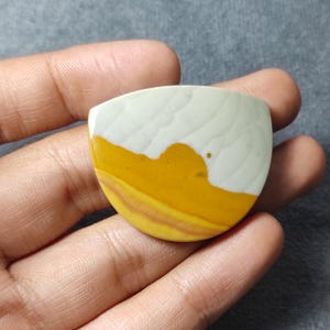 May include: A semi-circular cabochon stone featuring a smooth, polished surface with a distinct color split. The top section is a pale, creamy off-white, while the bottom half displays vibrant bands of mustard yellow and warm, earthy tan tones.