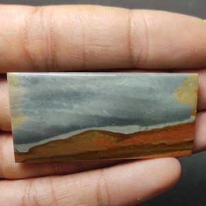 May include: A rectangular polished gemstone cabochon featuring horizontal bands of color. The stone displays a smooth, glossy surface with a palette of muted blue-gray, a thin white line, and earthy tones of ochre, brown, and reddish-orange.
