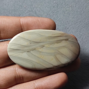 Könnte beinhalten: Ein ovaler, polierter Edelstein-Cabochon mit einer glatten Oberfläche und fließenden, geschichteten Mustern in Creme-, Beige- und sanften Grautönen. Der Stein hat eine glänzende Oberfläche und zeigt natürliche, organische Maserungen.
