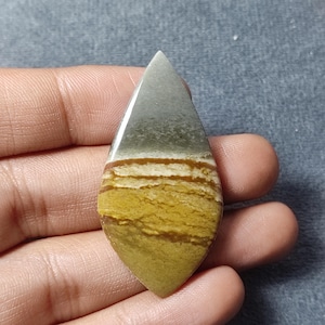 May include: A teardrop-shaped polished gemstone cabochon featuring horizontal bands of color. The top section is a smooth muted gray, transitioning into distinct layers of dark brown, cream, and a textured mustard yellow at the base.