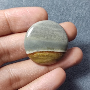 May include: A circular, polished cabochon stone featuring a smooth, glossy surface. The stone displays a distinct horizontal color gradient, transitioning from a muted gray upper section to a vibrant, earthy mustard yellow base.