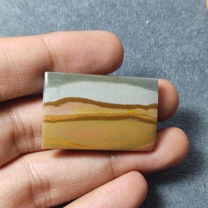 May include: A rectangular polished gemstone cabochon featuring horizontal bands of color including muted olive green, light gray, dark brown, golden yellow, and soft orange, creating a layered landscape-like appearance with a smooth surface.