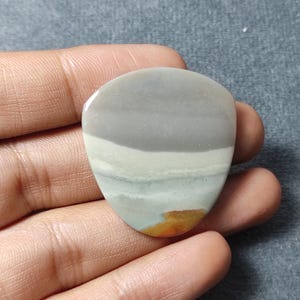May include: A polished, triangular-shaped stone cabochon featuring horizontal bands of color. The stone displays a smooth, glossy surface with layers of light gray, off-white, and pale green, accented by a small, distinct patch of warm orange-brown.