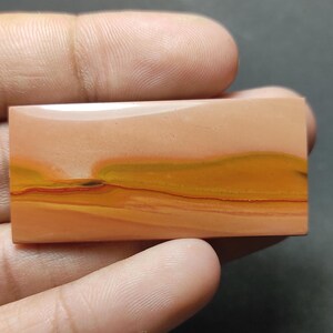 May include: A rectangular polished gemstone cabochon featuring horizontal bands of color in shades of peach, mustard yellow, and deep orange. The stone has a smooth, glossy surface with a distinct layered pattern resembling a landscape horizon.