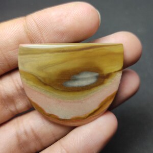 May include: A semi-circular polished gemstone cabochon featuring horizontal bands of mustard yellow, tan, and soft pink, with a central horizontal streak of grey. The stone has a smooth, glossy surface and a flat top edge with a rounded bottom.