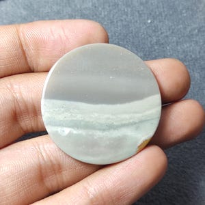 May include: A circular, flat stone cabochon featuring horizontal bands of muted gray, pale sage green, and off-white. The surface is smooth and polished, displaying natural mineral layering with a small touch of golden-brown color at the edge.