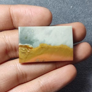 May include: A rectangular polished gemstone cabochon featuring a smooth surface with distinct horizontal color bands. The top section is a pale greyish-blue, transitioning into a vibrant mustard yellow and a warm peach-toned base layer.