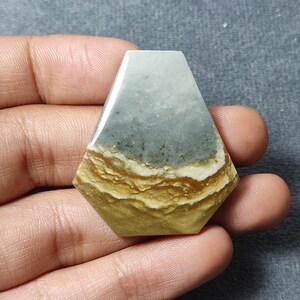 May include: A polished, pentagonal-shaped gemstone cabochon featuring a smooth surface with distinct horizontal color bands. The stone displays a top section of muted grey, transitioning into layered, wavy stripes of white, tan, and golden brown.