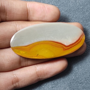 May include: An oval-shaped polished gemstone cabochon featuring a smooth, matte light gray upper section and a vibrant, banded lower section with swirling layers of mustard yellow, deep orange, and thin streaks of dark red throughout the surface.