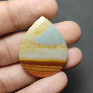 May include: A teardrop-shaped polished gemstone featuring horizontal bands of color including light blue, cream, white, golden yellow, and deep orange-brown. The stone has a smooth, glossy surface with distinct natural mineral layers visible throughout.
