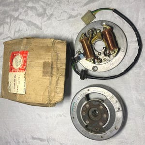 以下が含まれることがあります： 銅コイルを備えたホンダのオートバイ用ステーターアセンブリと円形の金属製ローター。31100-350-004のテキストとホンダのロゴが入った赤と白のラベルが付いた、使い古された段ボール箱が添えられており、すべて一緒に表示されています。