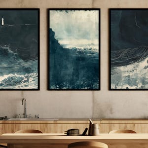May include: A set of three framed vertical wall art prints featuring dark blue and white ocean scenes. The outer prints display a small white lighthouse amidst crashing waves, while the centre print depicts a stormy sea and rocky coastline.