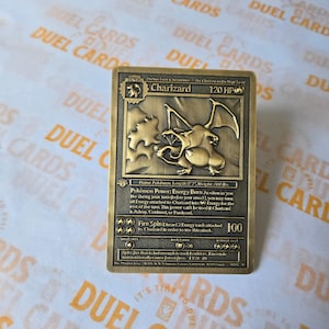 May include: A rectangular gold-toned metal trading card featuring a raised relief design of the Charizard character. The card displays the name Charizard, 120 HP, and detailed text including Energy Burn and Fire Spin abilities with a 100 damage value.