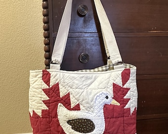 May include: A quilted tote bag featuring a white goose with a dark brown wing and yellow beak centered on a red and white geometric background. The bag has two off-white fabric shoulder straps attached with metal rings and a checkered interior lining.