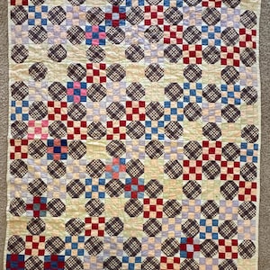 May include: A rectangular quilt featuring a repeating patchwork pattern of dark brown plaid octagons interspersed with smaller squares in solid red, light blue, pink, and pale yellow, all arranged on a cream-colored background with visible stitching.