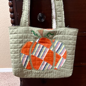 May include: A quilted sage green tote bag featuring a large central pumpkin appliqué. The pumpkin is constructed from alternating patches of solid orange fabric and multi-colored striped fabric, finished with a brown stem and green vines.