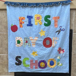 Könnte beinhalten: Ein hellblaues Stoffbanner mit weißen Punkten und einem dunkelblauen Rüschenrand oben. Das Banner zeigt bunte Stoffbuchstaben mit der Aufschrift FIRST DAY OF SCHOOL, verziert mit Applikationen eines roten Apfels, gelben Sternen, einer Schere und einem Herz.