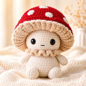 May include: A crocheted mushroom plush toy featuring a bright red cap with white spots and a beige underside. The figure has a cream-coloured body, two large black plastic eyes, a small stitched mouth, and a ruffled collar around the neck.