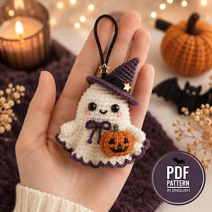 May include: A small crocheted ghost ornament in cream yarn with a purple witch hat, a tiny orange pumpkin, and a black hanging loop. The ghost features black bead eyes, a stitched smile, and pink cheeks. A circular badge reads PDF PATTERN IN ENGLISH.