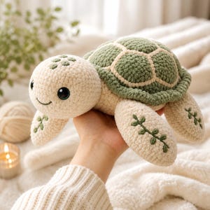May include: A crochet turtle plush toy featuring a cream-coloured head and limbs with dark green embroidered leaf details. The shell is textured in a deep sage green with cream-coloured borders. The turtle has large black plastic eyes and a stitched smile.