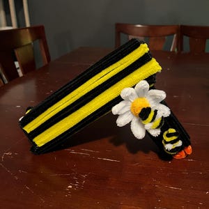 May include: A decorative craft item constructed from black and yellow pipe cleaners, featuring a striped rectangular base and a prominent white daisy flower with a yellow center, accented by a small bee figure with black and yellow stripes.