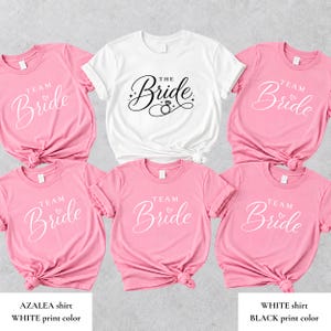 Op de afbeelding: Vijf roze t-shirts met witte tekst Team Bride en één wit t-shirt met zwarte tekst The Bride inclusief een ringafbeelding. De t-shirts hebben korte mouwen en een ronde hals, waarbij de roze shirts zijn voorzien van een klein hart-icoon.