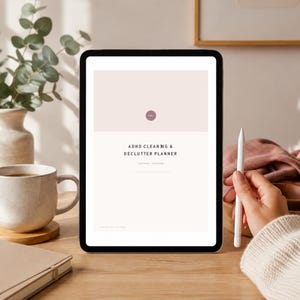 Può includere: Un tablet che mostra un pianificatore digitale ADHD Cleaning & Declutter con un'intestazione rosa pallido, una piccola icona circolare e testo nero. Lo schermo mostra il titolo e il sottotitolo, con uno stilo bianco posizionato accanto.