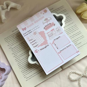 Pode incluir: Uma ficha de leitura de papel retangular com um padrão xadrez rosa claro e branco. A página inclui seções rotuladas como Book Review, Start Date, Finish Date, Rating, Title, Author, Genre, Pages, Tropes, Thoughts e Favorite Quote.