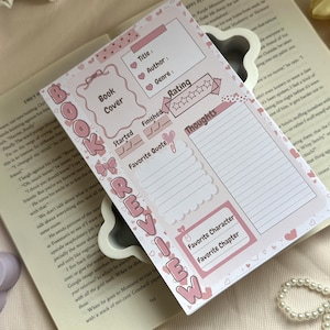 Peut inclure: Une fiche de lecture rectangulaire en papier avec un design rose clair et blanc. La page comporte des sections pour le titre, l'auteur, le genre, la note, les dates de début et de fin, la citation préférée, les pensées, le personnage et le chapitre favoris.