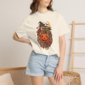 May include: A cream-colored short-sleeve t-shirt featuring a graphic print of a snake wearing a witch hat coiled around a jack-o'-lantern pumpkin. The illustration includes autumn leaves, a crescent moon, and small bats in shades of orange and black.
