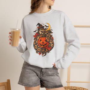 May include: A light gray crewneck sweatshirt featuring a graphic print of a snake wearing a witch hat coiled around a jack-o'-lantern pumpkin. The design includes autumn leaves, a crescent moon, and small bats, set against a heathered fabric background.