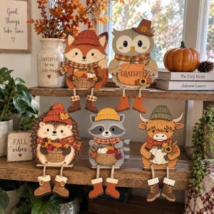 May include: A set of five wooden shelf sitters featuring a fox, owl, hedgehog, raccoon, and highland cow, each wearing a hat and scarf, holding a basket or pumpkin, and dangling boots. The owl holds a pumpkin with the word GRATEFUL printed on it.