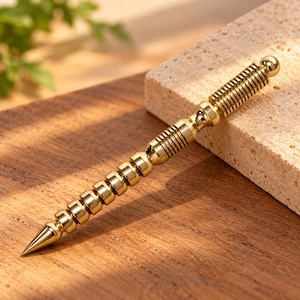 May include: A gold-colored metal tool with a pointed tip and a cylindrical body featuring a series of stacked, rounded segments and a ribbed grip section. The object has a smooth, reflective finish and a small spherical knob at the end.