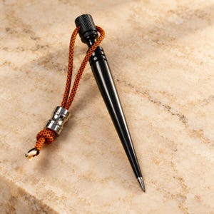 May include: A black metal marlinspike tool featuring a tapered, pointed tip and a knurled top. An orange and black patterned paracord lanyard is attached to the top, secured by a silver-colored metal bead and a knot at the end of the cord.