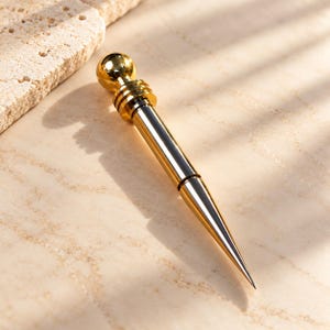 May include: A metallic gold-toned pointed tool featuring a spherical top and a series of three horizontal rings below the sphere. The body of the tool is cylindrical and tapers to a sharp, fine point at the bottom end, with a polished finish.